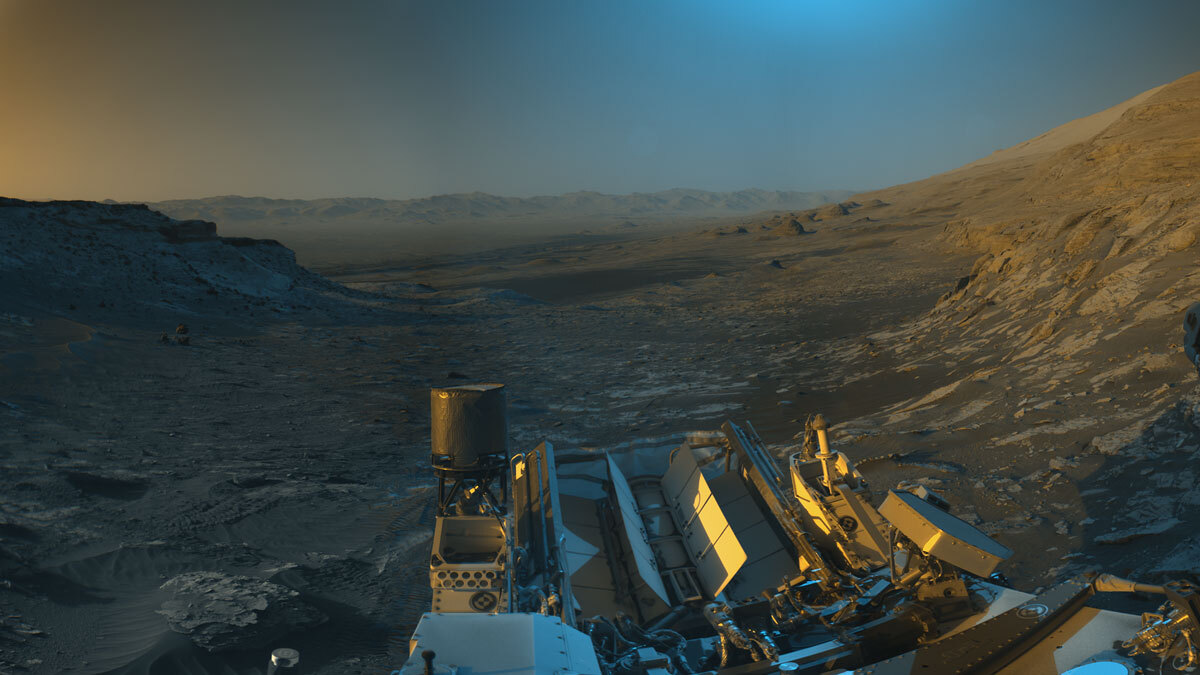 Марсоход Curiosity НАСА использовал свои черно-белые навигационные камеры для съемки панорам этой сцены в два времени суток; синий, оранжевый и зеленый цвета были добавлены к комбинации обеих панорам для художественной интерпретации сцены. 