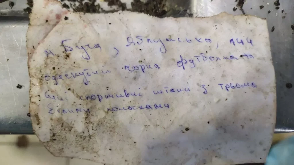 Новые доказательства того, как российские солдаты казнили жителей города Буча 18 Записка, сфотографированная рядом с неопознанным телом и распространенная в Telegram, содержала подробную информацию об этом человеке для тех, кто может его искать. Позже этот человек был опознан как Валера Котенко. Записка гласила: "Город Буча, улица Яблонская, 144. Одет в черную футболку и синие спортивные штаны с тремя белыми"