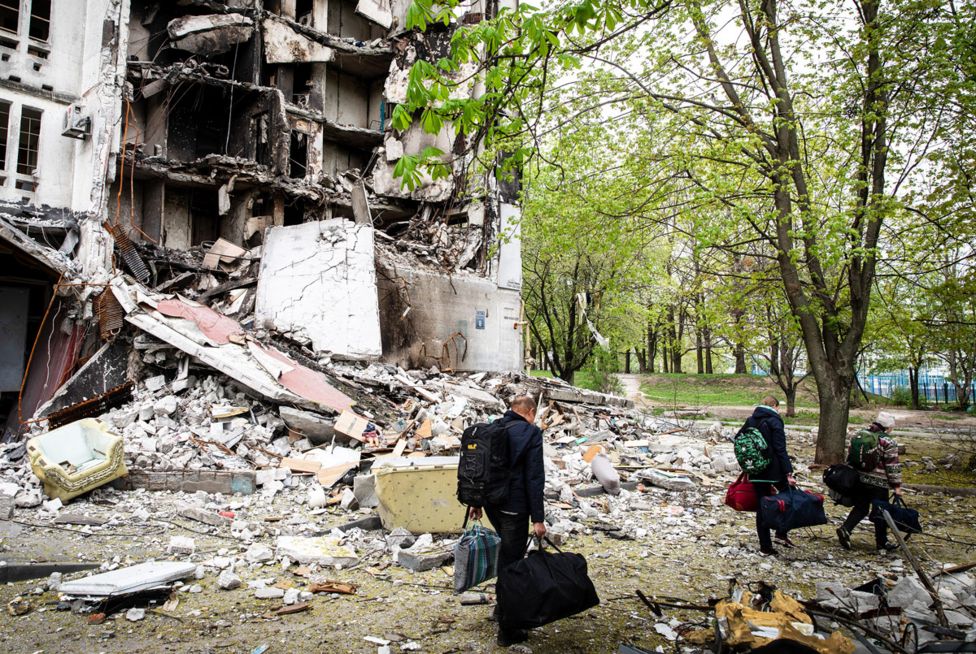 Жители с собранными сумками проходят мимо разрушенного здания в Салтовке, 29 апреля