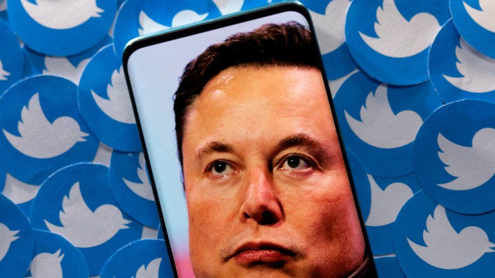 Elon Musk on phone screen with Twitter logos in the background.