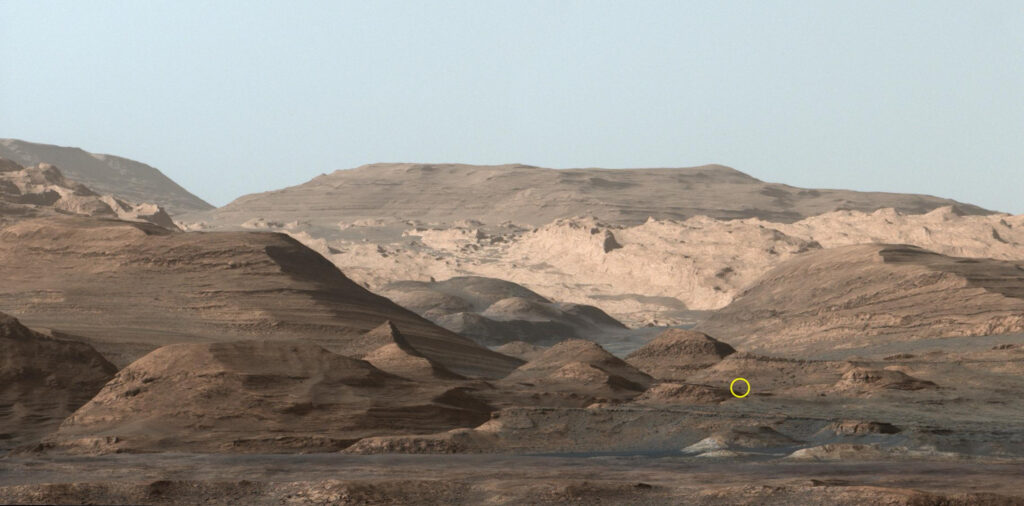 Спустя 10 лет после посадки марсоход NASA Curiosity все еще сохраняет драйв 2 Перевал Парайтепуй с расстояния: Эта сцена была запечатлена Curiosity 9 сентября 2015 года, когда марсоход НАСА находился за много миль от своего нынешнего местоположения. Круг указывает на местоположение валуна размером с Кьюриосити, мимо которого недавно проехал марсоход. Слева от него находится "Перевал Парайтепуй", через который Curiosity сейчас проезжает.
