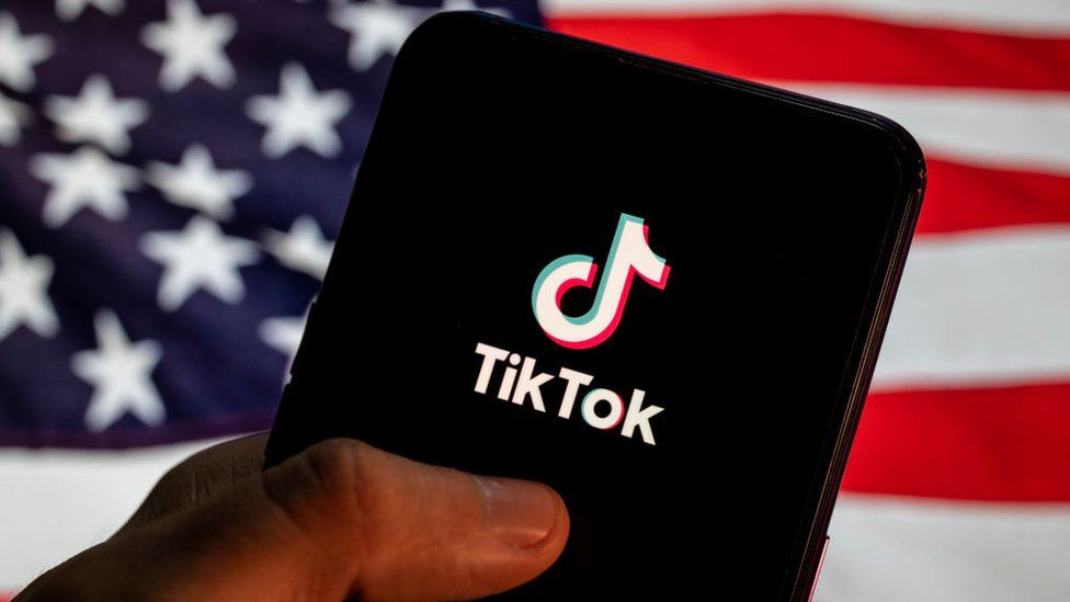 Логотип TikTok изображен на мобильном устройстве с флагом США на заднем плане.