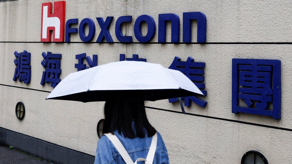 A woman carrying an umbrella walks past the logo of Foxconn outside a company building.