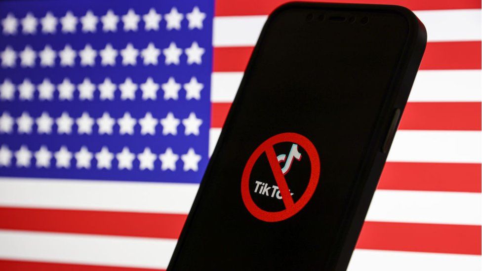 The TikTok logo is displayed on a mobile phone screen in front of the US flag.