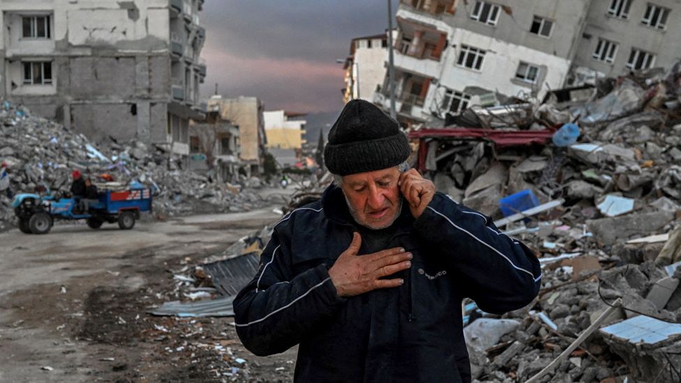 A man speaks on the phone in front of collapsed buildings in Samandag, Turkey
