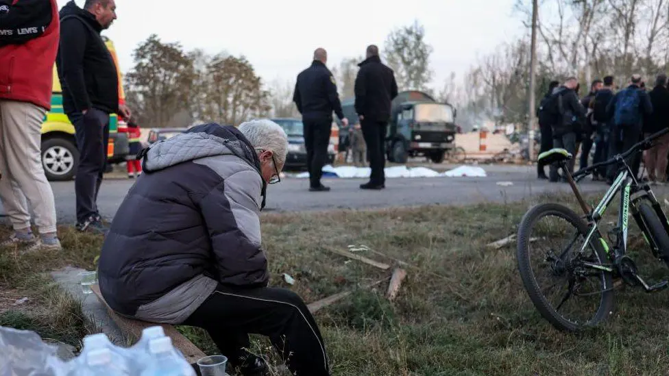 Местные жители на месте военного удара в селе Гроза на северо-востоке Украины, 5 октября 2023 года. На переднем плане виден мужчина, сидящий на бетонном выступе среди клочья травы.