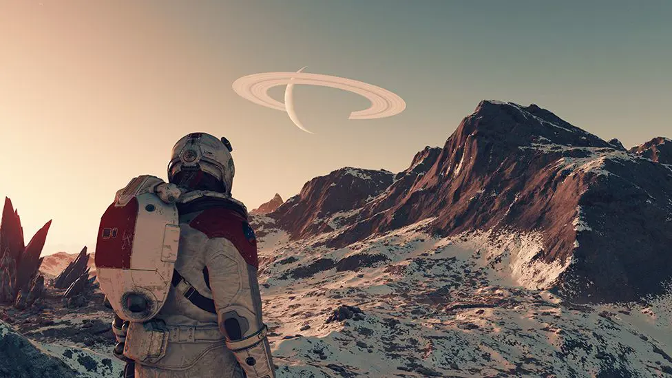 На снимке экрана из Starfield изображена спина астронавта в скафандре и шлеме, осматривающего бесплодный ландшафт планеты. Он скалистый, с выступающими из земли скалами. Скалы покрыты белым порошком – то ли пылью, то ли снегом. Вдалеке на небе виднеется еще одна планета и ее горизонтальные кольца.