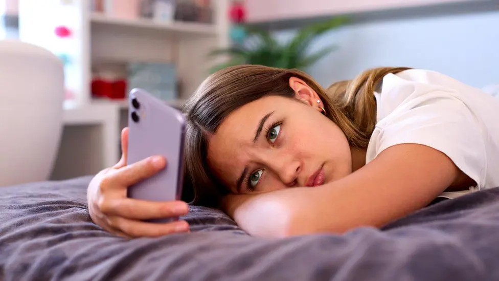 Девочка-подросток лежит дома на кровати и смотрит на мобильный телефон