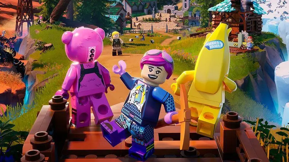 Персонаж LEGO с улыбающимся и дерзким выражением лица стоит между ярко-розовым медведем Lego и желтым гуманоидным бананом — все трое созданы по образцу игровых персонажей из Fortnite. Они находятся на ярко окрашенном поле, а за ними простирается сцена: овцы из LEGO, пасущиеся на траве, и видны постройки, включая церковь, замок и бревенчатую хижину. Там происходит много активности - мы видим, как персонажи рубят деревья и слоняются по деревне вдалеке.