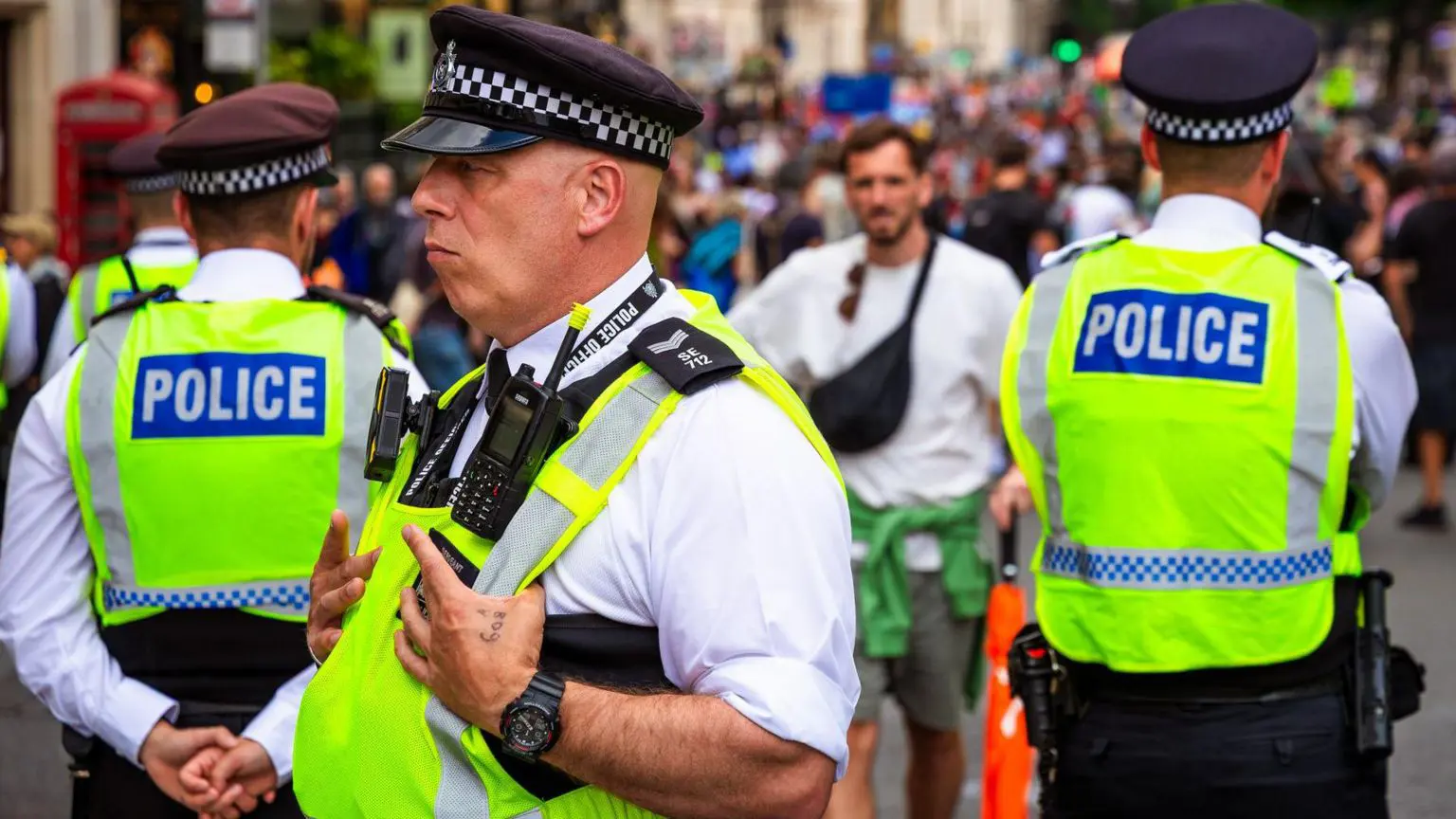 Регулятор по правам человека критикует использование Лондонской полицией камер распознавания лиц 1 Трое полицейских в светоотражающих жилетах стоят в толпе людей, когда к ним подходит мужчина в летней одежде.