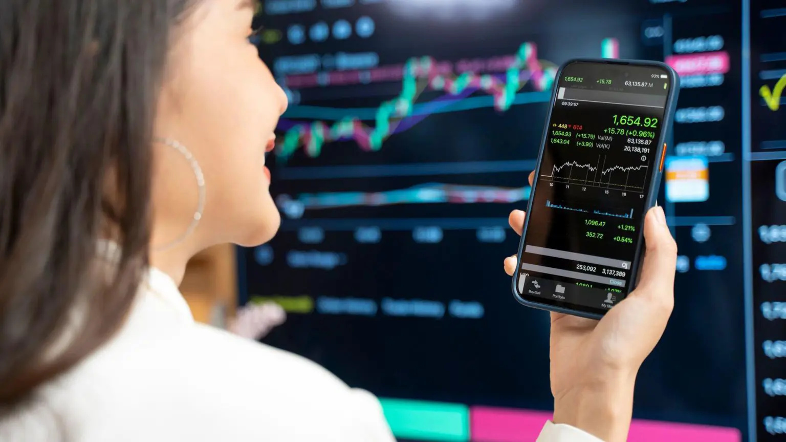 Пользователей криптовалюты заставляют делиться данными своих счетов с налоговыми органами 1 Женщина в белой рубашке смотрит на линейный график на своем телефоне, на фоне большого черного экрана, отображающего биржевые торги.