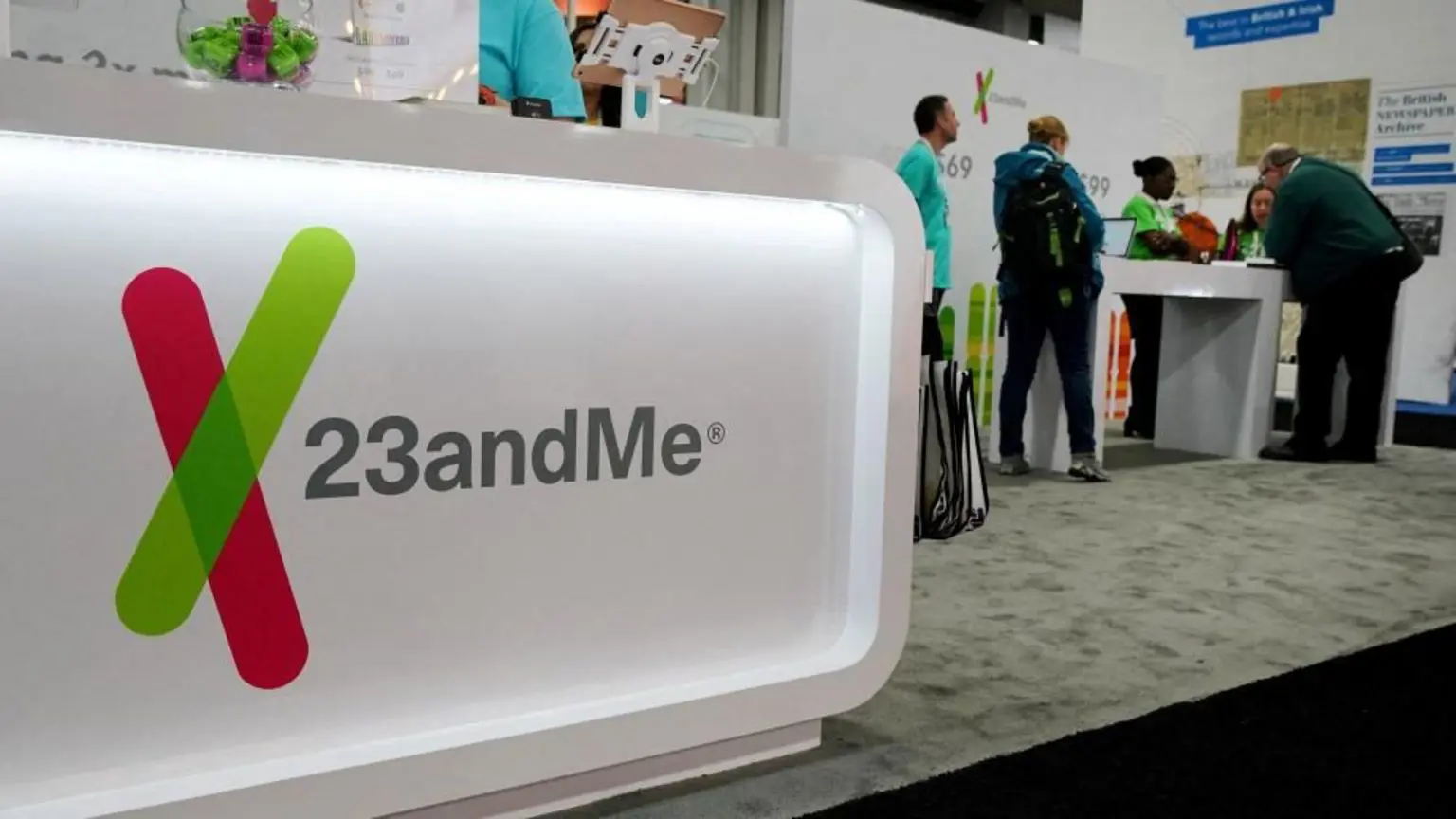 Генеральный прокурор Калифорнии подал в суд на компанию-преемника 23andMe за утечку данных в 2023 году. 1 Логотип 23andMe можно увидеть на генеалогической конференции. Логотип расположен на ярко-белом стенде в передней части выставочного павильона компании, покрытого ковром. На заднем плане за столом сидят люди в светло-голубых форменных рубашках и другие в повседневной одежде.