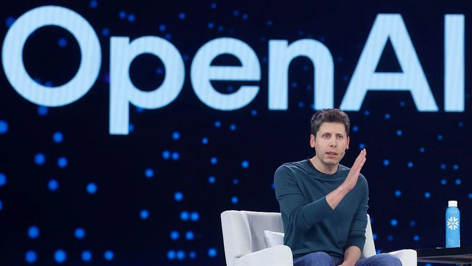 Компания OpenAI планирует выйти на биржу, что усиливает конкуренцию за инвестиции с компанией Anthropic. 1 Сэм Альтман сидит на стуле на сцене. Он жестикулирует руками. Позади него — логотип Open AI.