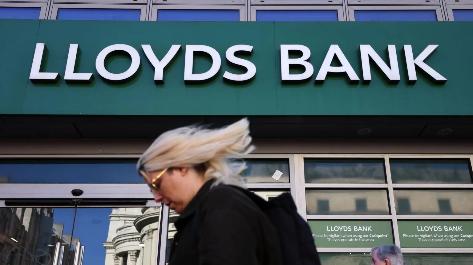 Пользователи приложений Lloyds, Halifax и Bank of Scotland столкнулись с перебоями в работе. 1 Логотип банка Lloyds на фасаде банка на главной улице. На переднем плане проходит женщина со светлыми волосами.