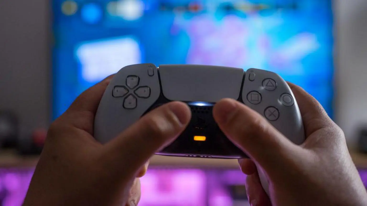 Сеть PlayStation Network недоступна, тысячи геймеров не могут играть. 1 Белый контроллер PlayStation 5, который кто-то держит в руках на фоне размытого экрана телевизора.