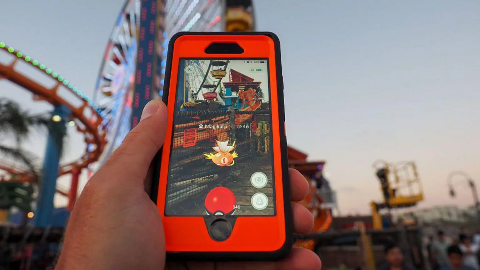 10 лет Pokémon Go, и миллионы всё ещё пытаются поймать их всех. 3 Красный и черный телефон, на экране которого отображается изображение локации с улучшенным Магикарпом из Pokémon Go. На размытом фоне видны колесо обозрения и трасса американских горок.